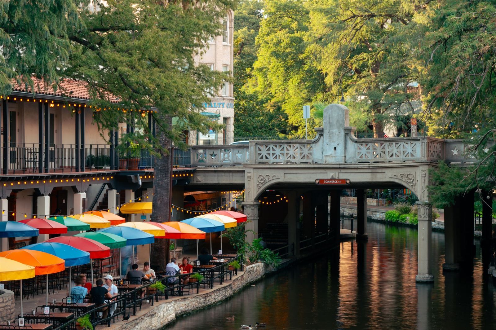 San Antonio River Walk with colorful umbrellas over cafe tables beside the river and an ornate stone bridge
