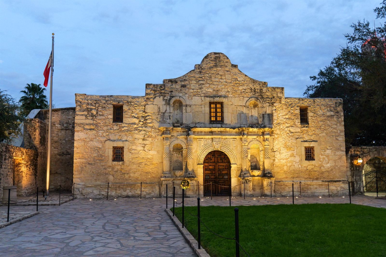 The stone facade of the Alamo church at dusk, with a flag flying beside it