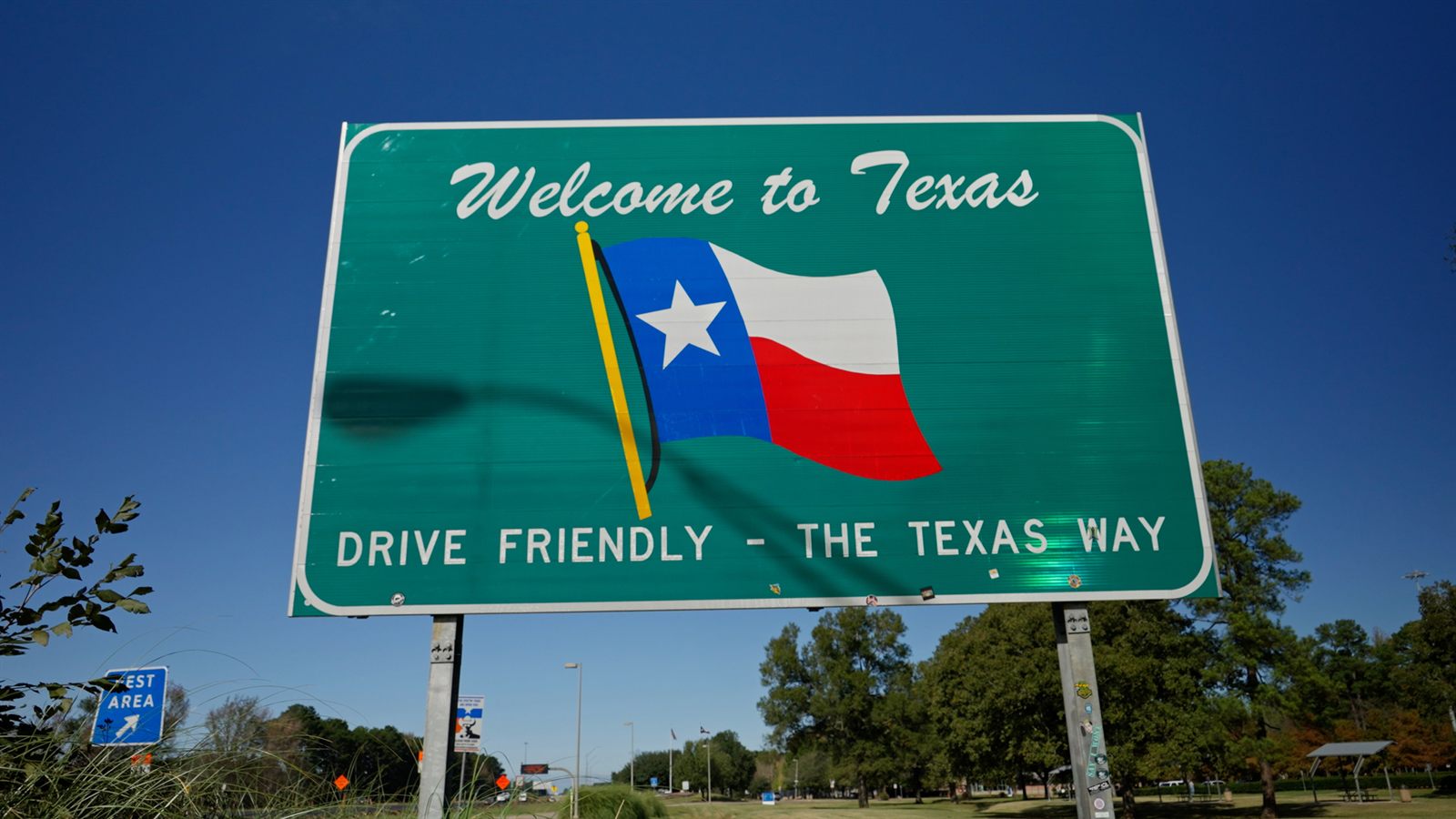 A green roadside sign reading Welcome to Texas, Drive Friendly, The Texas Way, with a Texas flag